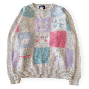 May include: A light grey sweater with a patchwork design. The sweater features squares of pastel colours, including pink, blue, and purple, each with a different floral or abstract pattern. The sweater has a ribbed hem and cuffs.