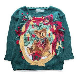 Peut inclure: Pull vert en tricot avec un motif festif représentant un ours en peluche, des nœuds rouges et des éléments décoratifs. Le pull a un col rond et des manches longues, parfait pour la saison des fêtes.