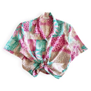 May include: A short-sleeved shirt with a collar and tie-front. The shirt displays a patchwork design with pink, teal, and beige floral and geometric patterns. The fabric has a slightly textured appearance.