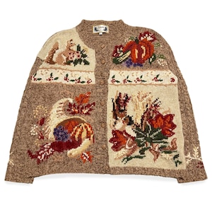 May include: A brown cardigan with embroidered autumn designs. The sweater has a round neckline, button closure, and detailed images of pumpkins, flowers, and a squirrel. The colour palette includes shades of brown, beige, red, and green.