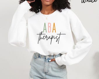 May include: White sweatshirt with the text "ABA therapist" in a script font. The letters "ABA" are in pastel colours. The word "White" is in the upper right corner.