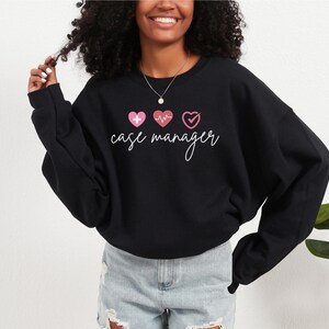 Könnte beinhalten: Schwarzes Sweatshirt mit dem Schriftzug "case manager" in Schreibschrift. Über dem Text befinden sich drei rosa Herzen, jedes mit einem anderen Design: ein Kreuz, ein Herzschlag und ein Häkchen.