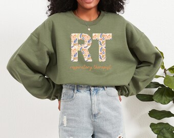 Może przedstawiać: Zielona bluza z literami "RT" w kwiatowy wzór. Poniżej pomarańczowym kolorem napisano "respiratory therapist". Bluza jest noszona z dżinsowymi szortami.