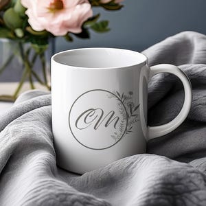 Op de afbeelding: Witte keramische mok met een handvat, met een grijs bloemenkransontwerp rond de letters "OM". De mok staat op een grijze stoffen ondergrond, met een vaas met roze bloemen op de achtergrond.