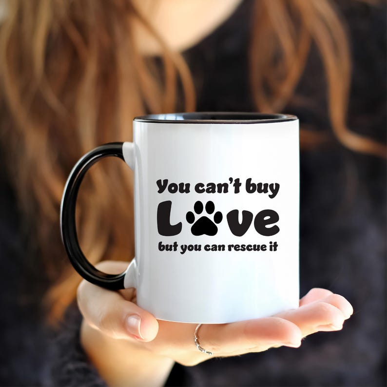 May include: White ceramic mug with a black rim and handle. The mug has a black paw print and the text "You can't buy love but you can rescue it".