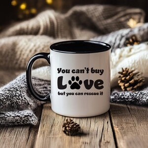 May include: White ceramic mug with a black rim and handle. The mug has a black paw print and the text "You can't buy love but you can rescue it".
