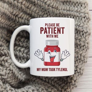 May include: White ceramic mug with the text "PLEASE BE PATIENT WITH ME" in maroon. Below the text is a cartoon pill bottle with a smiling face and the text "MY MOM TOOK TYLENOL". The mug has a large, curved handle.