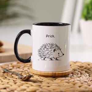 K&ouml;nnte beinhalten: Wei&szlig;e Keramiktasse mit schwarzem Henkel und Rand. Die Tasse zeigt eine schwarz-wei&szlig;e Illustration eines Igels und das Wort "Prick." &uuml;ber dem Igel. Die Tasse steht auf einem Holzuntersetzer.