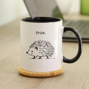 K&ouml;nnte beinhalten: Wei&szlig;e Keramik-Tasse mit schwarzem Innenraum und Henkel. Die Tasse zeigt eine schwarz-wei&szlig;e Illustration eines Igels und das Wort "Prick". Die Tasse steht auf einem runden Untersetzer aus Holz.