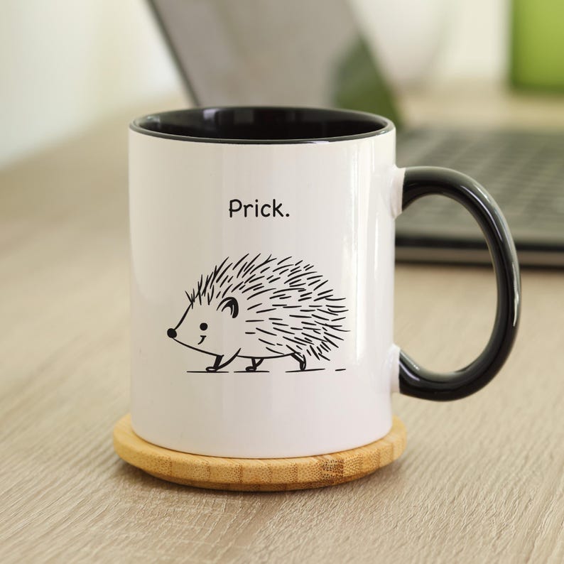 K&ouml;nnte beinhalten: Wei&szlig;e Keramik-Tasse mit schwarzem Innenraum und Henkel. Die Tasse zeigt eine schwarz-wei&szlig;e Illustration eines Igels und das Wort "Prick". Die Tasse steht auf einem runden Untersetzer aus Holz.