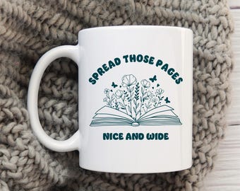 Könnte beinhalten: Weiße Keramik-Tasse mit dem Text "SPREAD THOSE PAGES NICE AND WIDE" in Türkis. Das Design zeigt ein offenes Buch mit Blumen und Schmetterlingen. Die Tasse steht auf einer grauen Strickoberfläche.