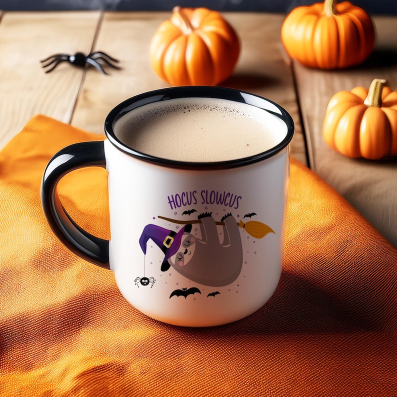 May include: A white ceramic mug with a black handle and rim, filled with a light brown beverage. The mug features a cartoon sloth wearing a witch hat and holding a broom, with the text "Hocus Slowcus". Halloween-themed with pumpkins and a spider.
