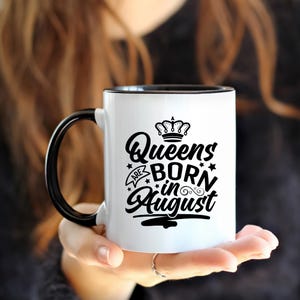 May include: White ceramic mug with a black handle and interior. The mug features the text "Queens are born in August" in black script, along with a crown and star accents. A ring is visible on the person's finger holding the mug.