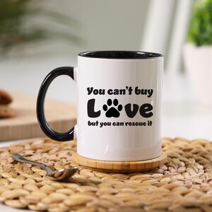 May include: White ceramic mug with a black rim and handle. The mug has a black paw print and the text "You can't buy LOVE but you can rescue it".