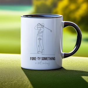 May include: White ceramic mug with a black rim and handle. The mug features a line drawing of a golfer mid-swing and the text "FORE-TY SOMETHING & still sucking" in black. The mug is on a green surface.