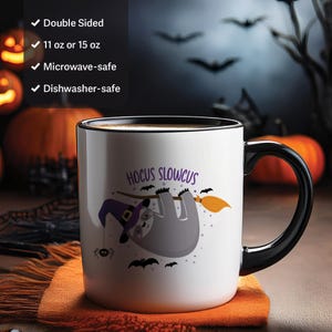 May include: White ceramic mug with a black handle and rim, featuring a Halloween-themed sloth illustration. The sloth is wearing a witch hat and hanging from a branch with a broom. The mug has the text "Hocus Slowcus" and is double-sided.