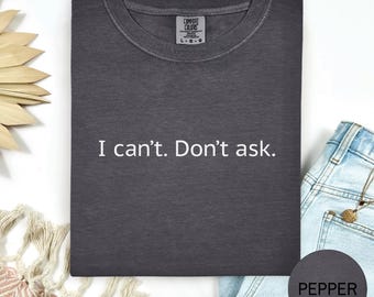 May include: A dark gray Comfort Colors t-shirt with the text "I can't. Don't ask." in white. The shirt is folded and displayed with a pair of blue jeans and a decorative element.