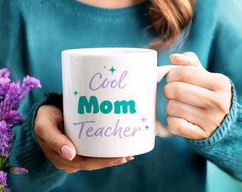 Puede incluir: Taza de cerámica blanca con las palabras "Cool Mom Teacher" en escritura turquesa y lavanda. La taza tiene un asa y la sostiene una persona que lleva un jersey turquesa. La taza está decorada con estrellas.
