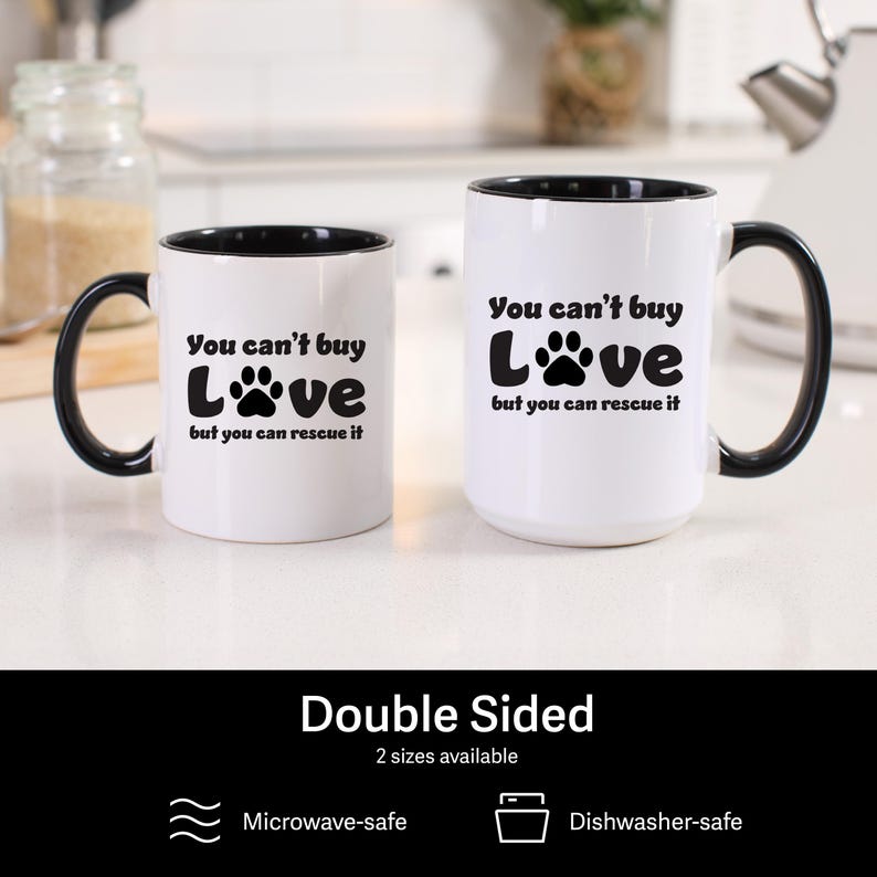 May include: Two white ceramic coffee mugs with black handles and black rims. The mugs have a paw print design and the text "You can't buy Love but you can rescue it".