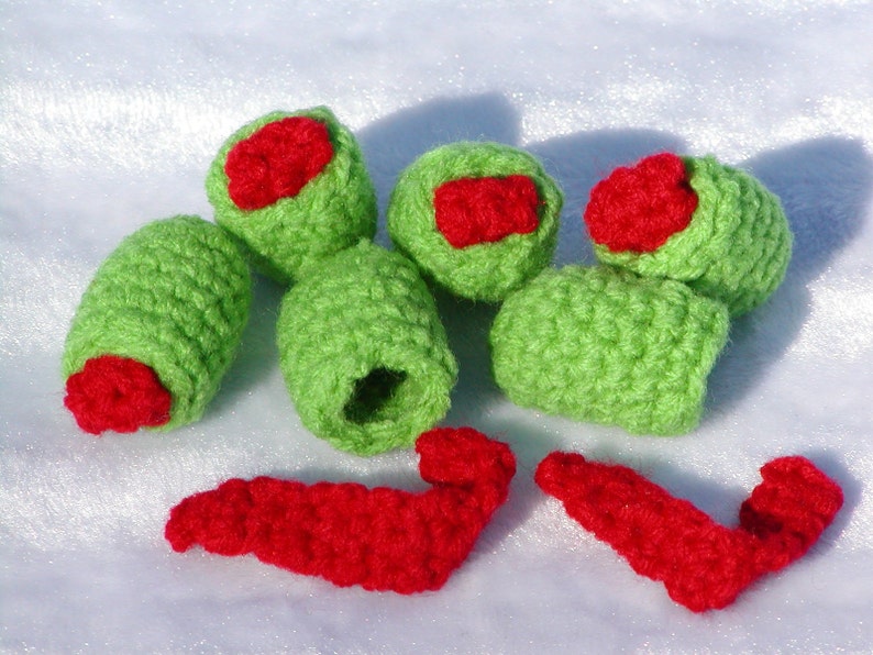 May include: Six crocheted green olives with red pepper filling. The olives are arranged in a cluster on a white surface. There are also three red crocheted pepper pieces.
