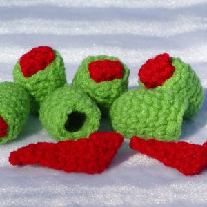 May include: A set of five crocheted green olives with red pepper filling. The olives are arranged in a semi-circle with two red pepper pieces in front.