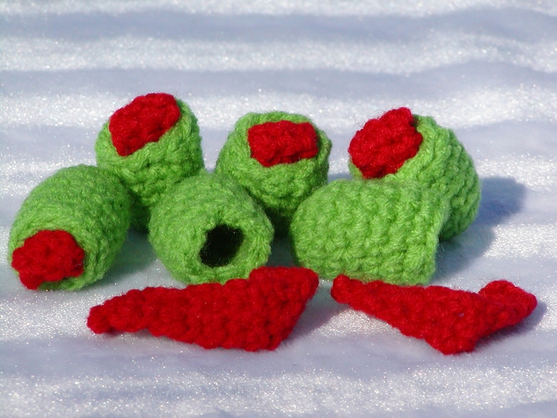 May include: A set of five crocheted green olives with red pepper filling. The olives are arranged in a semi-circle with two red pepper pieces in front.