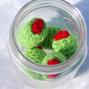 May include: A glass jar filled with crocheted green olives with red centers. The olives are arranged in a circular pattern.