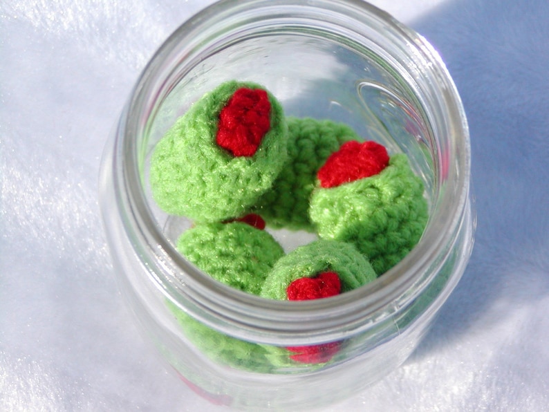 May include: A glass jar filled with crocheted green olives with red centers. The olives are arranged in a circular pattern.