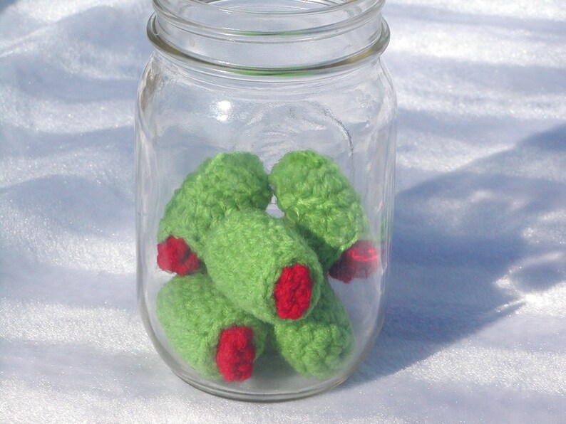 May include: A glass jar filled with crocheted green and red olives. The olives are handmade and have a realistic appearance.