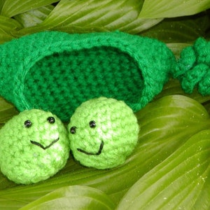 May include: Two green crocheted peas with smiling faces inside a green crocheted pea pod. The pod is lying on green leaves.