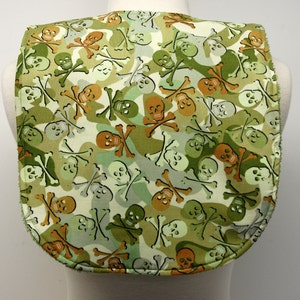 May include: A green and white fabric bib with a skull and crossbones pattern. The bib has a scalloped edge and is made of a soft, comfortable material.