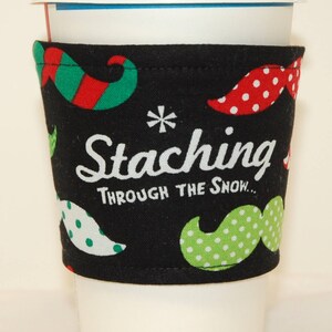 May include: A black coffee cup sleeve with a festive mustache pattern in red, green, and white polka dots. The text "Staching Through the Snow..." is printed in white. The sleeve is wrapped around a white paper coffee cup.