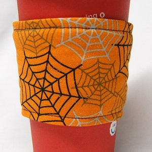 May include: A red paper coffee cup with a white lid, featuring an orange fabric sleeve. The sleeve is patterned with black and silver spiderwebs, ideal for Halloween. The cup is designed for hot drinks.