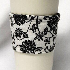 May include: A white to-go coffee cup with a black and white floral fabric sleeve. The sleeve has a repeating pattern of black flowers and vines on a white background.