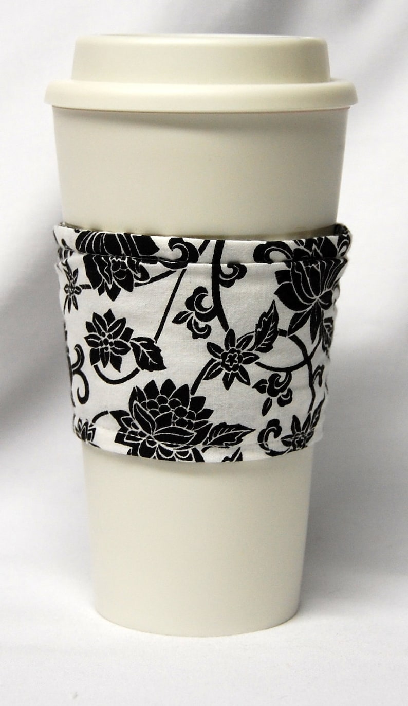 May include: A white to-go coffee cup with a black and white floral fabric sleeve. The sleeve has a repeating pattern of black flowers and vines on a white background.