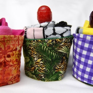 May include: Three fabric covered containers with different patterns. The containers are filled with socks and topped with a decorative ball. The containers are orange with a floral pattern, green with a tropical leaf pattern, and purple with a checkered pattern.