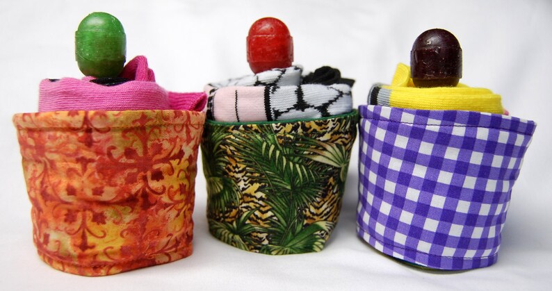 May include: Three fabric covered containers with different patterns. The containers are filled with socks and topped with a decorative ball. The containers are orange with a floral pattern, green with a tropical leaf pattern, and purple with a checkered pattern.
