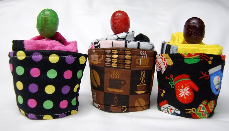 May include: Three colorful fabric coffee cup cozies with different patterns. The cozies are made from socks and feature a polka dot pattern, a coffee cup pattern, and a Christmas mitten pattern.