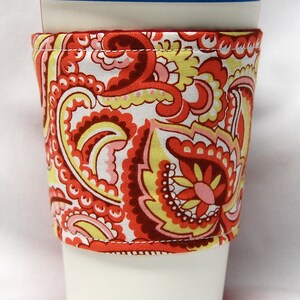 Puede incluir: Una taza de café blanca con una funda de tela de color rojo, amarillo y rosa con estampado paisley. La funda está cosida con hilo rojo.