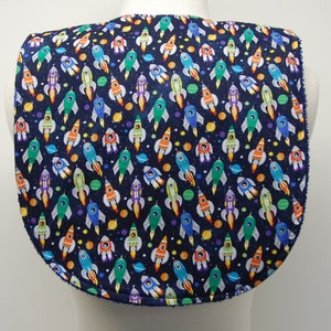 Puede incluir: Un babero azul oscuro con un patrón de cohetes y planetas coloridos. El babero tiene un escote redondeado y un borde festoneado.