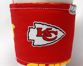 May include: A red and yellow fabric coffee cup sleeve with a white arrowhead design and the letters "KC" in red.