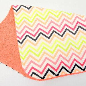 May include: A baby burp cloth featuring a coral terry cloth backing and a chevron pattern front in black, pink, orange, and neon yellow. The cloth is rectangular with rounded corners.