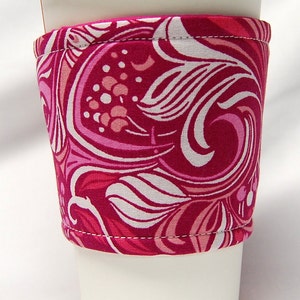May include: A reusable coffee cup sleeve with a vibrant floral pattern in shades of pink and white. The fabric sleeve wraps around a white paper coffee cup, providing insulation and a stylish look. The sleeve is designed to fit standard coffee cup sizes.