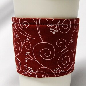 May include: A white travel mug with a cream-colored lid and a burgundy fabric sleeve. The sleeve features a white floral and swirl pattern. The mug is designed for hot beverages.