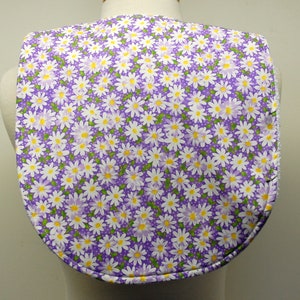May include: Purple fabric bib with a white daisy pattern. The bib has a rounded neckline and a scalloped edge.