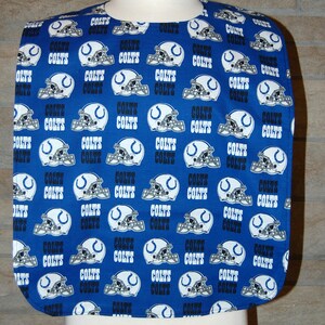Puede incluir: Un babero de tela azul y blanco con un patrón repetido de logotipos de los Indianapolis Colts. El babero tiene un escote redondeado y una parte inferior ligeramente puntiaguda.