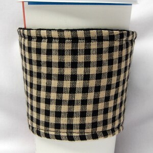 May include: A white paper coffee cup with a white lid, and a black and beige checked fabric coffee sleeve. The sleeve is wrapped around the cup, providing insulation and a stylish look. The cup is on a white background.