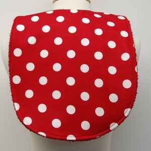 May include: Red fabric bib with white polka dots. The bib has a rounded neckline and a white trim around the edges.