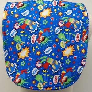May include: Blue fabric bib with a superhero theme. The bib features a repeating pattern of red and green superheroes with yellow stars and lightning bolts on a blue background. The words "SUPER", "HERO", and "DUPER" are printed in white speech bubbles.