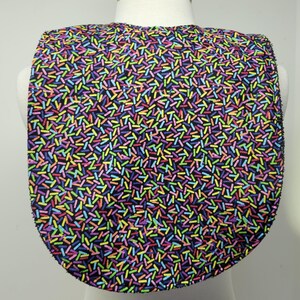 May include: A baby bib with a black background covered in a colorful pattern of small, irregular, vertical lines in shades of yellow, green, blue, purple, and pink. The bib is U-shaped and made of fabric.